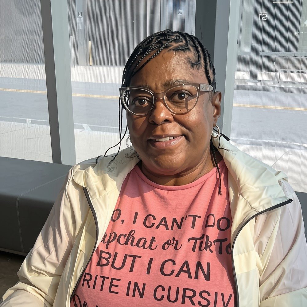 Black woman with hair pulled back. She is wearing glasses, a white jacket and a pink shirt with writing on it and sitting in front of a window that is facing the street.