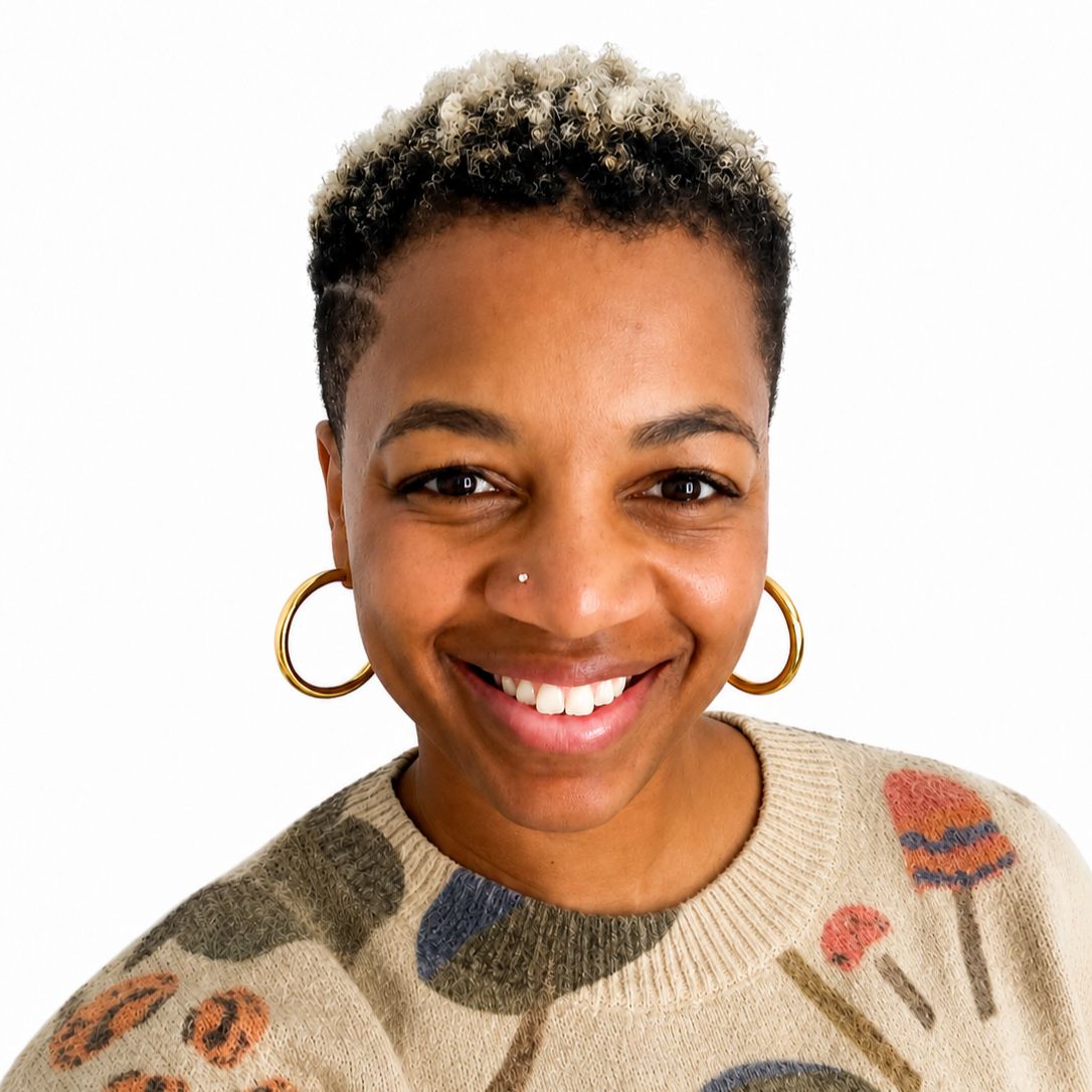 Black woman with short curly black hair with blonde tips wearing hoop earrings and a beige sweater with various colored shapes on it