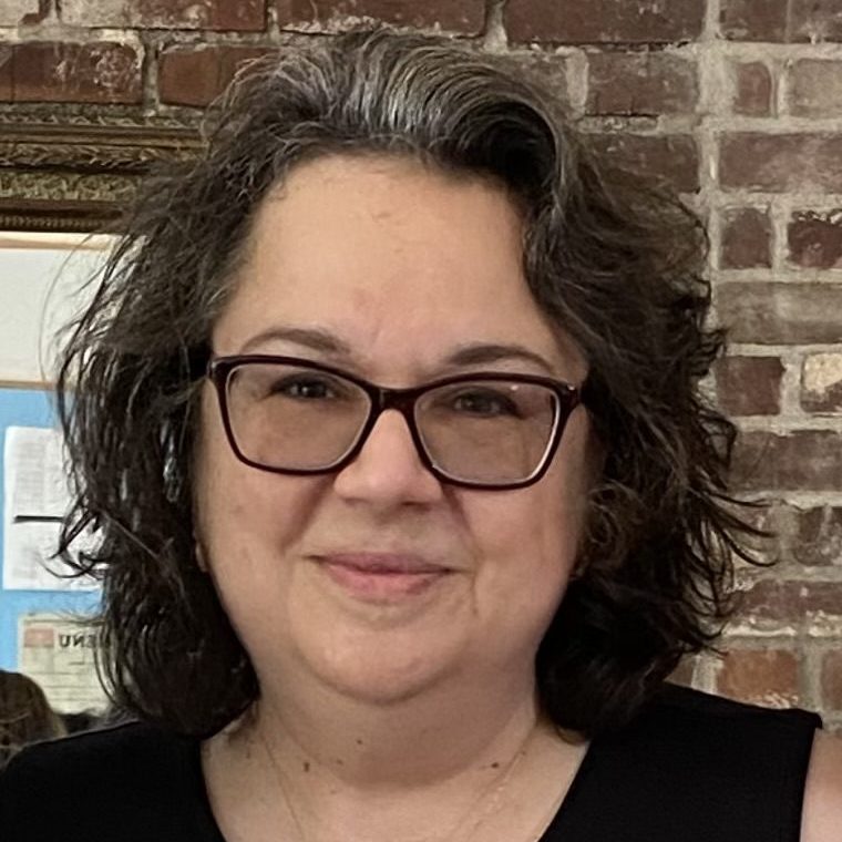White woman with curly short brown hair wearing glasses and a short sleeve black shirt.